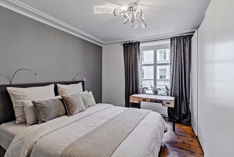 Modern bedroom with a large bed, nightstands, a small table, and curtains. The room features wooden flooring and a contemporary chandelier.