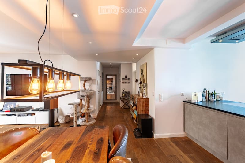 Open-plan living and dining area with wooden flooring, a large wooden dining table with chairs, a cat tree, a modern kitchen area with cabinets and appliances, and a hanging light fixture with exposed bulbs.