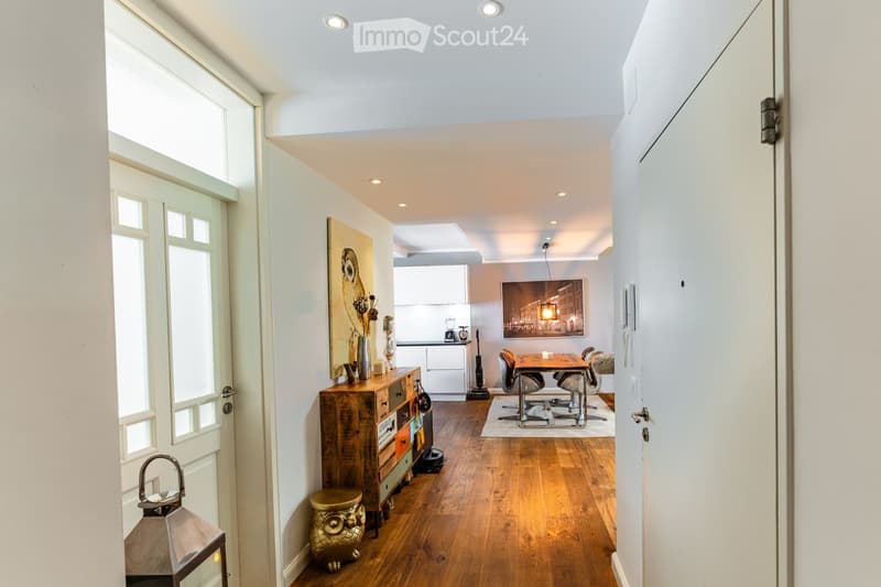 A well-lit, modern hallway leading into a dining area with a wooden dresser, table, and chairs. The space features wooden flooring, white walls, and contemporary decor including artwork and a lantern.