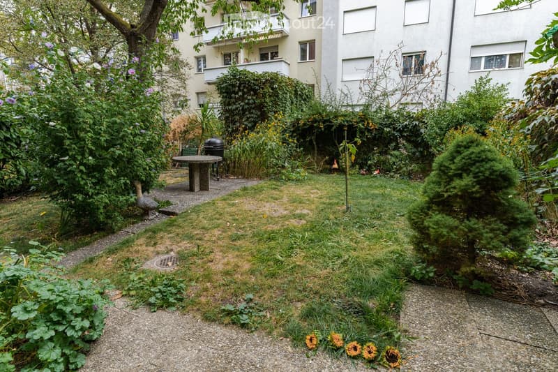 Garden with a mix of grass, gravel paths, and various plants, including sunflowers and shrubs. There is a round stone table, a grill, and a decorative bird statue. The garden is bordered by apartment buildings with balconies and windows.