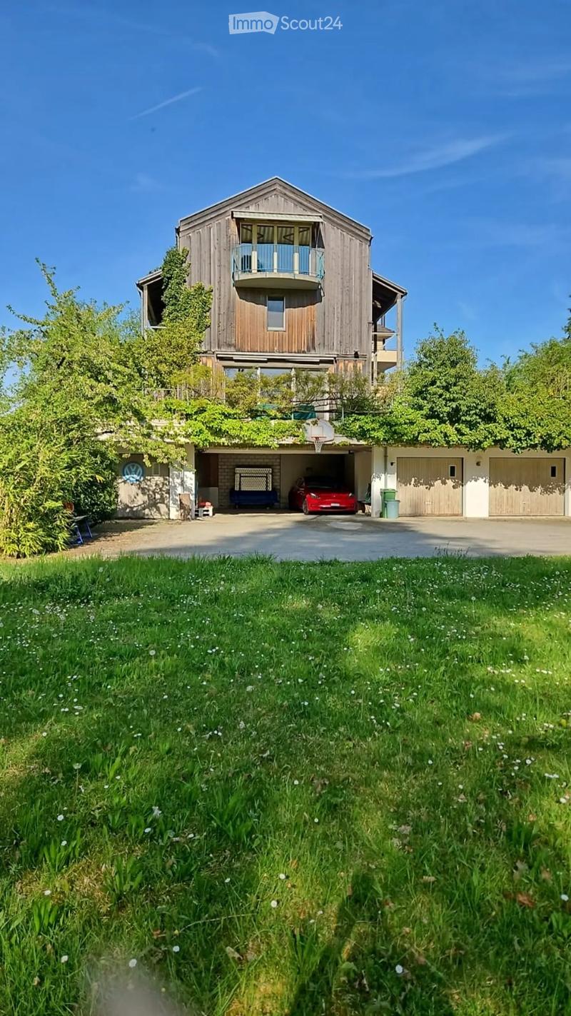 A two-story wooden house with a balcony, garage, and a basketball hoop. The house is surrounded by greenery and a well-maintained lawn.
