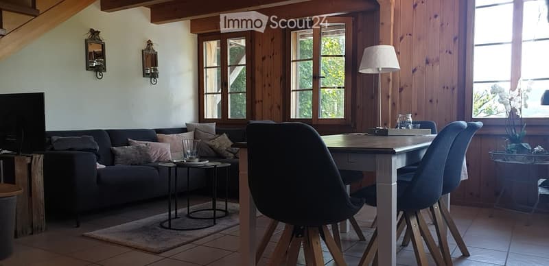 A cozy living room with wooden walls, large windows, a dark blue sofa with pillows, a small round table, a dining table with chairs, a floor lamp, and decorative mirrors on the wall.