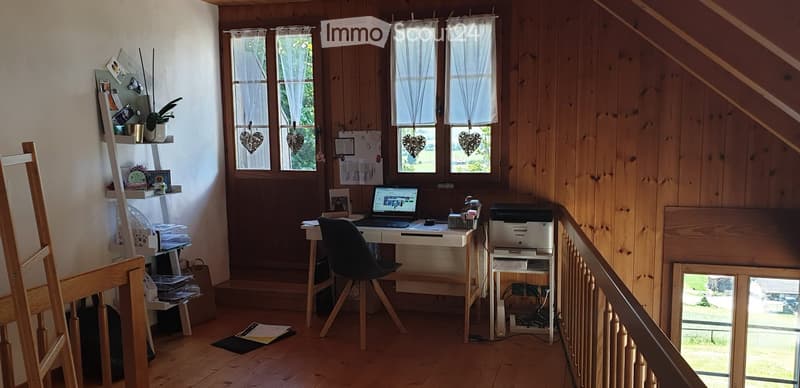 Cozy attic workspace with wooden walls, large windows, desk, chair, laptop, printer, shelf with books and plants, and a small stool.