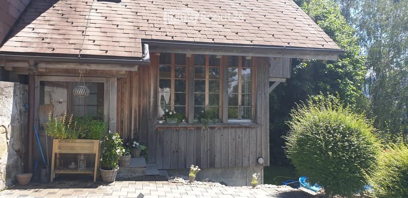 A rustic wooden house with a steeply pitched roof, large windows, and a small porch area decorated with potted plants and flowers. The house features a mix of wooden and stone elements, with a small garden area visible to the right.