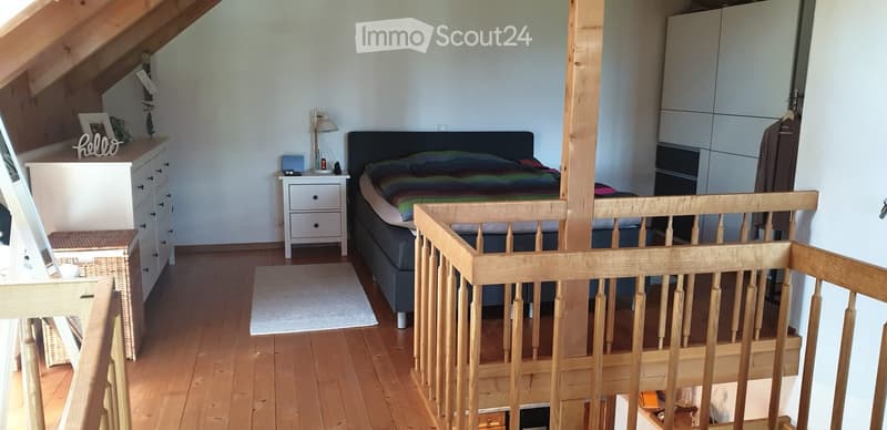 Cozy attic bedroom with wooden floors, slanted ceilings, a double bed with colorful bedding, a nightstand with a lamp, a wardrobe, and a small wooden railing in the foreground.