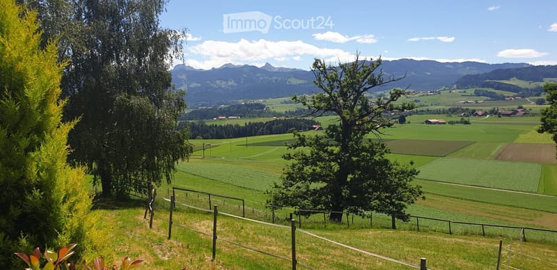 A panoramic view of a lush green valley with rolling hills, scattered farms, and distant mountains under a clear blue sky.