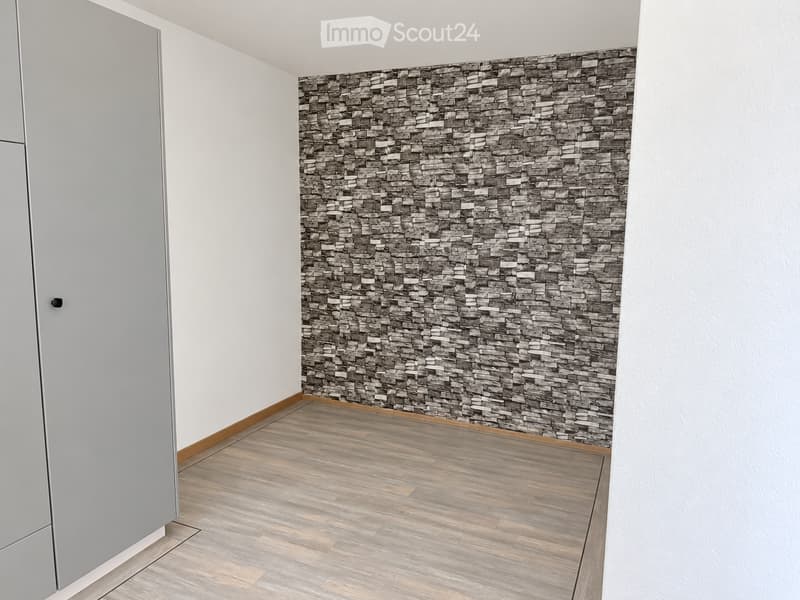 Empty room with light wooden flooring, white walls, and a decorative stone-patterned accent wall. A grey cabinet is visible on the left side.