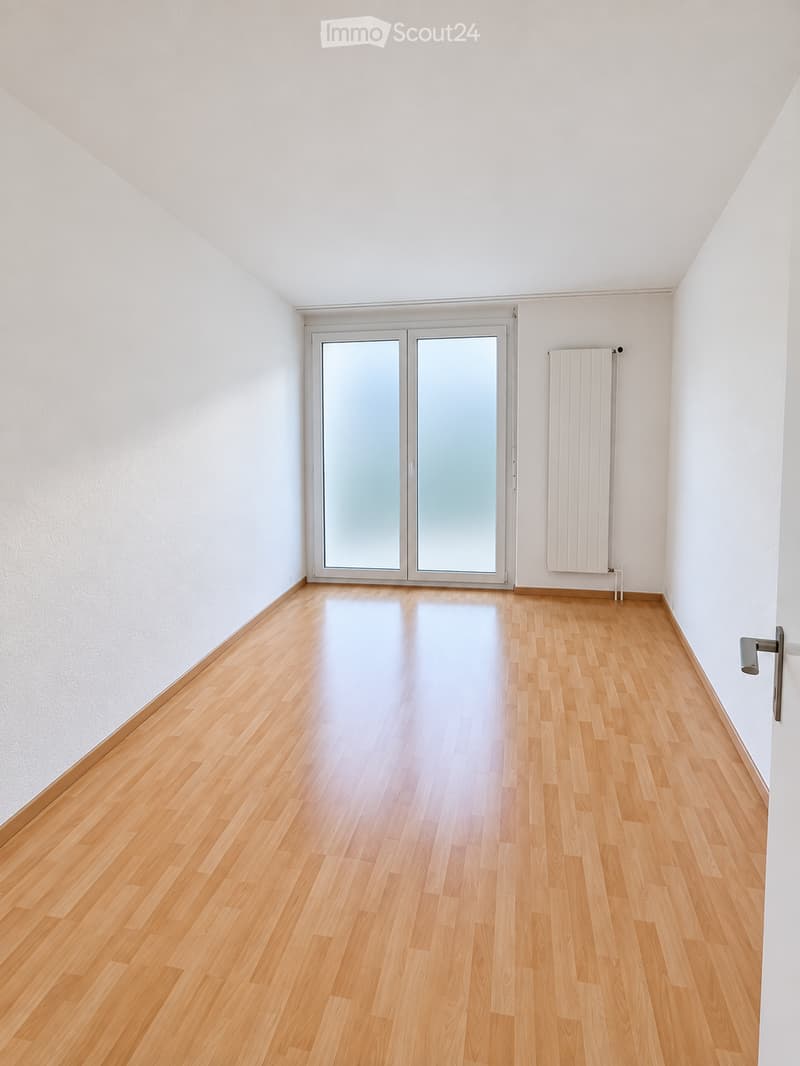 Empty room with light wooden flooring, white walls, and a large glass door leading outside.