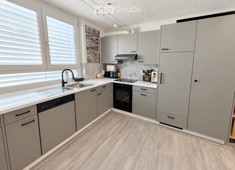 Modern kitchen with light grey cabinets, white countertops, and a light wood floor. Features include a sink with a black faucet, an oven, a dishwasher, and a coffee maker on the counter.