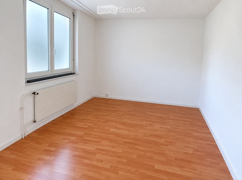 Empty room with light wooden flooring, white walls, and a large window with a radiator below it.