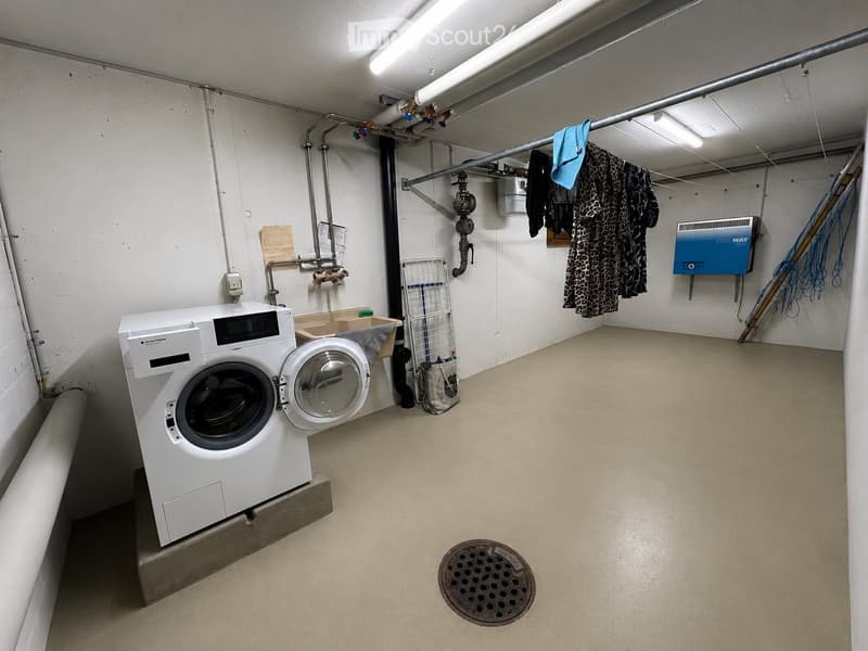 Laundry room with a washing machine and dryer, a laundry basket, a laundry rack, and a control panel. The room has exposed pipes and a drain in the floor.