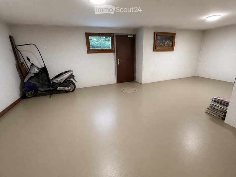 Empty room with white walls, a door, a window, and a scooter parked against the wall. There is a stack of magazines in the corner and a small wooden shelf with logs above the door.