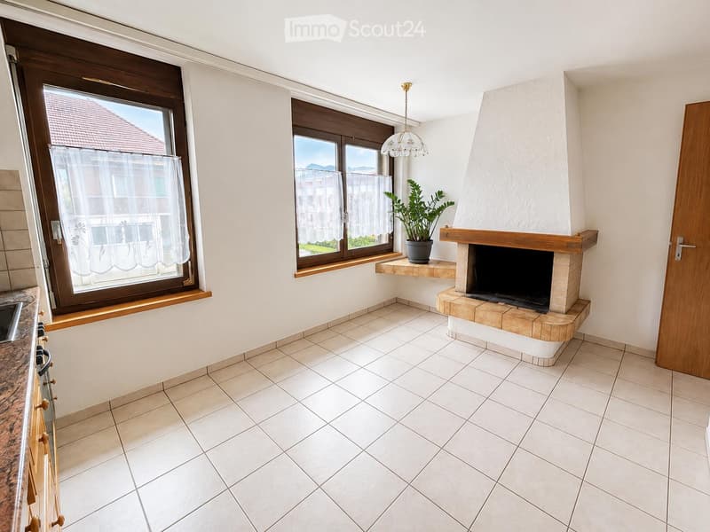 Bright living room with tiled flooring, large windows with white curtains, a central fireplace with a wooden mantel, and a hanging chandelier.