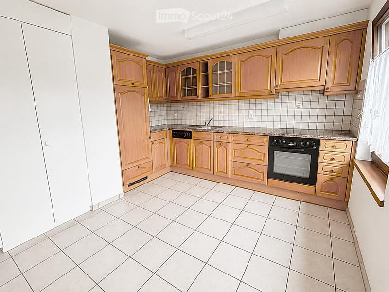 Bright kitchen with light wooden cabinets, tiled walls, and a white tiled floor. Features a sink, oven, and stove.