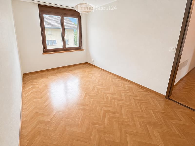 Empty room with light wooden herringbone flooring, white walls, and a large window with brown frame. The room has a pendant light fixture hanging from the ceiling.