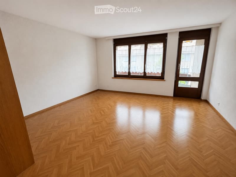 Empty room with wooden herringbone flooring, white walls, large window with brown frame, and a door with glass panels.