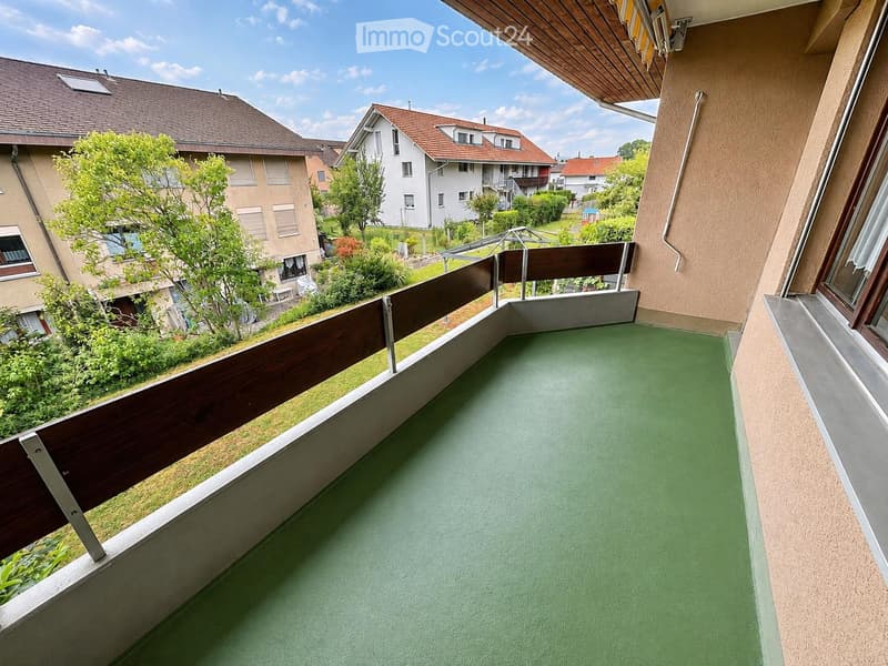 Balcony with green flooring, wooden railing, and a view of neighboring houses and greenery.