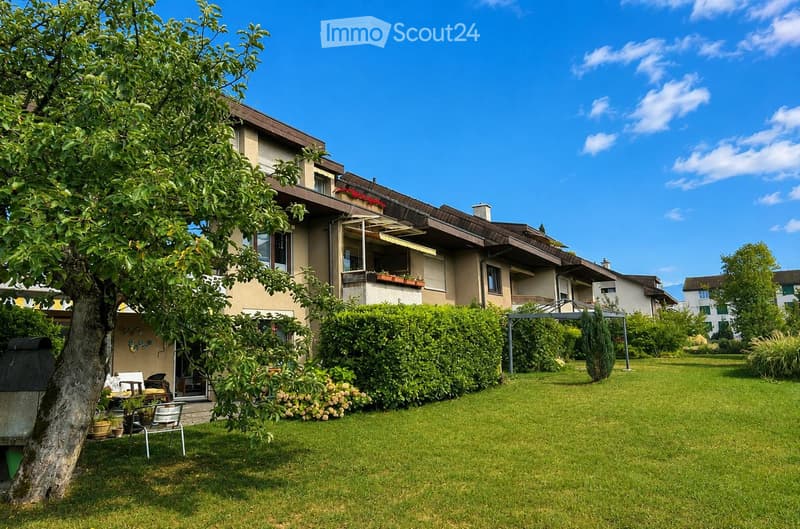 A well-maintained garden area with a lawn, a large tree, patio furniture, and a multi-story building with balconies and windows in the background.