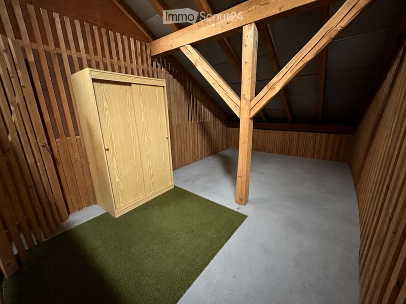 An empty attic space with wooden beams, exposed wooden walls, and a small wooden cabinet. The floor is partially covered with green carpet and concrete.
