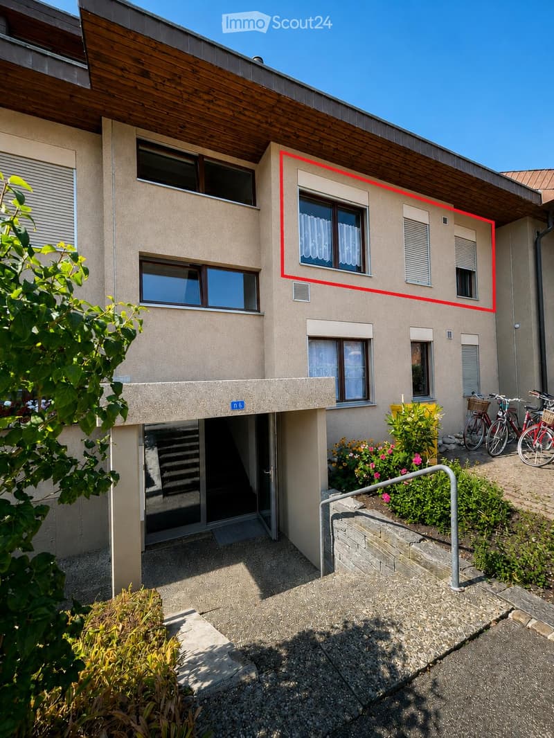 Modern two-story house with beige facade, large windows, and a wooden roof section. Features include a raised ground floor entrance, a small garden area with plants and flowers, and bicycles parked nearby.
