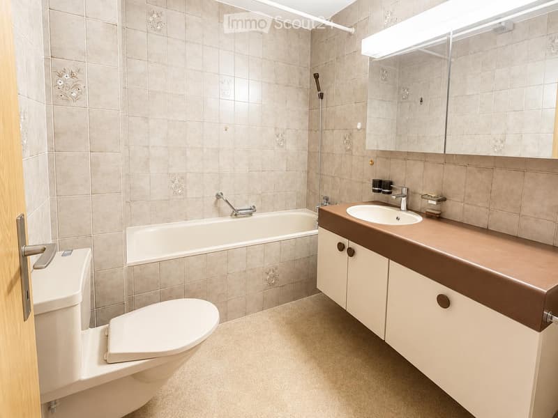 Modern bathroom with beige tiled walls, a bathtub, a toilet, a sink with a mirror, and a wooden cabinet.