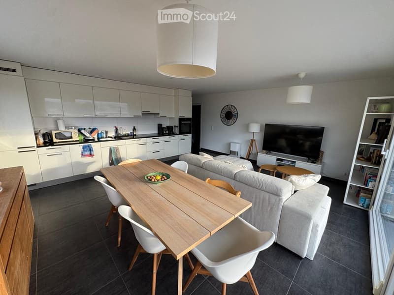 Open-plan living and dining area with a grey sofa, wooden dining table with white chairs, modern kitchen with white cabinets, microwave, and oven, and a television on a stand.