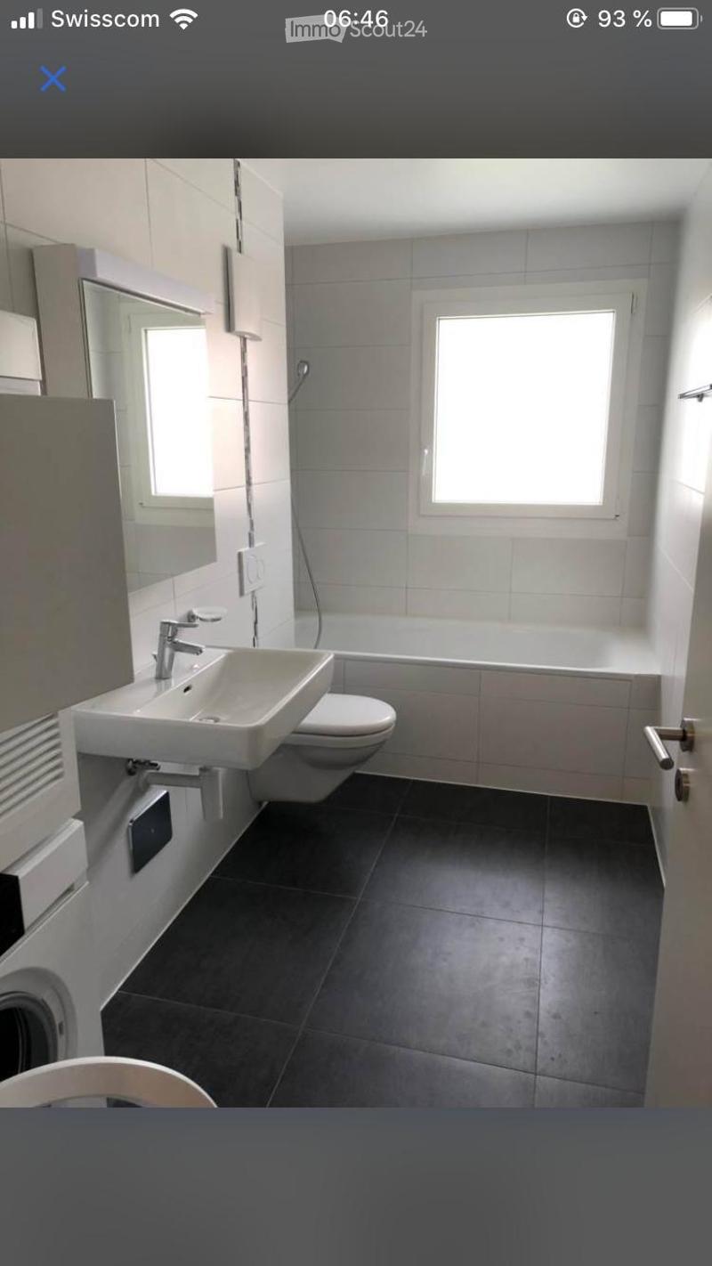 Modern bathroom with a white sink, toilet, bathtub, and large mirror. The room features dark tiled flooring and two windows providing natural light.