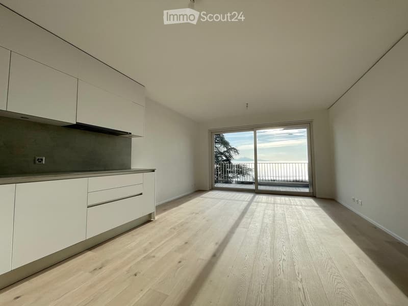 Empty kitchen with light wood flooring, white cabinetry, and a large window offering a scenic view of the sea and mountains.