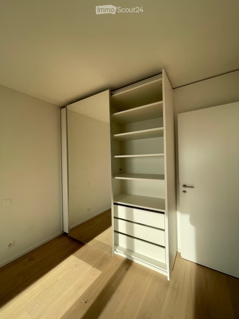 Empty room with light wooden flooring, white walls, and an open white built-in closet with shelves and drawers.