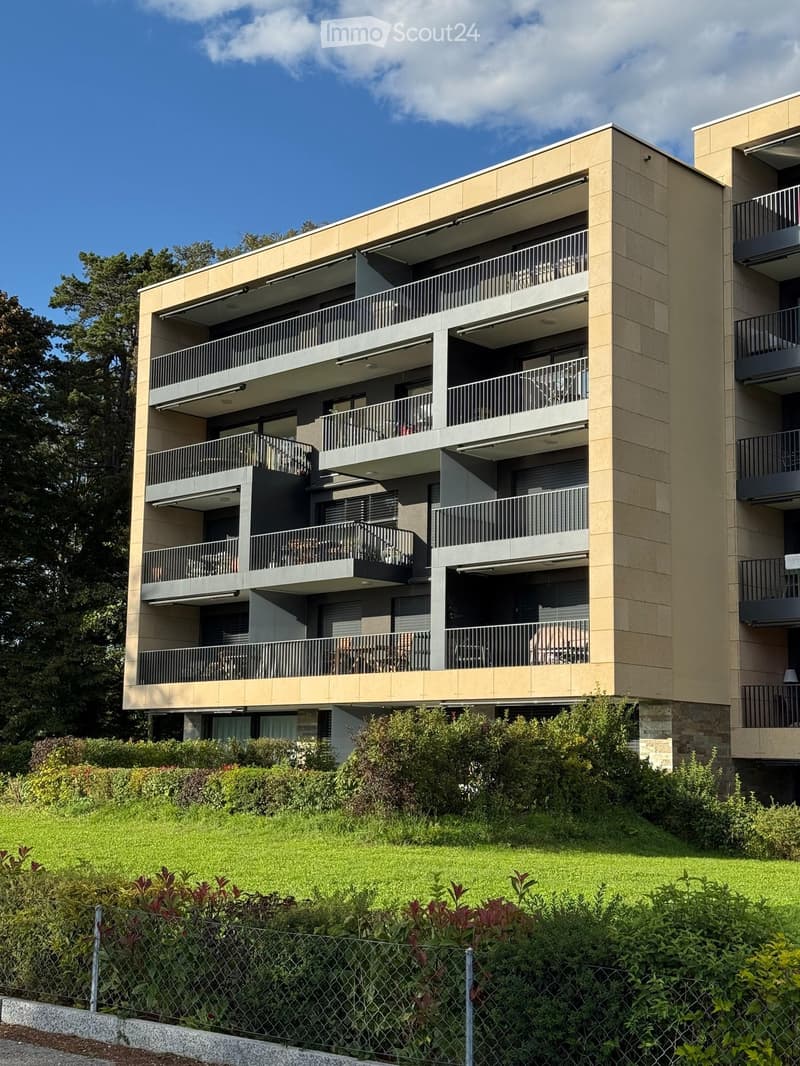 Modern multi-story apartment building with balconies, surrounded by greenery and a fence.