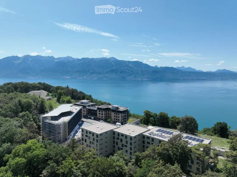 Aerial view of a modern building complex surrounded by trees, overlooking a lake and mountain range.