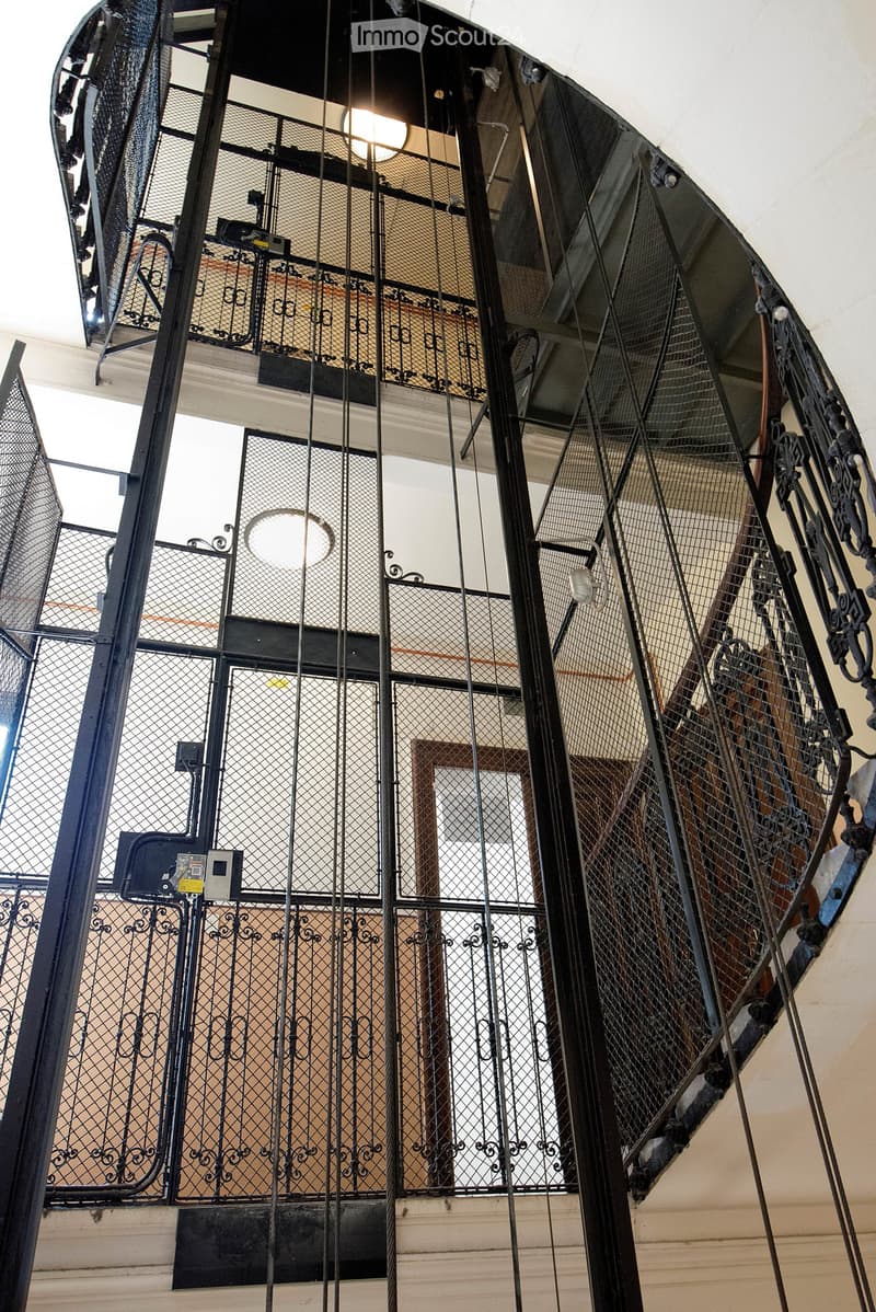 Ornate metal staircase with intricate wrought-iron railings and glass panels, leading to an upper level with visible wooden flooring and lighting fixtures.