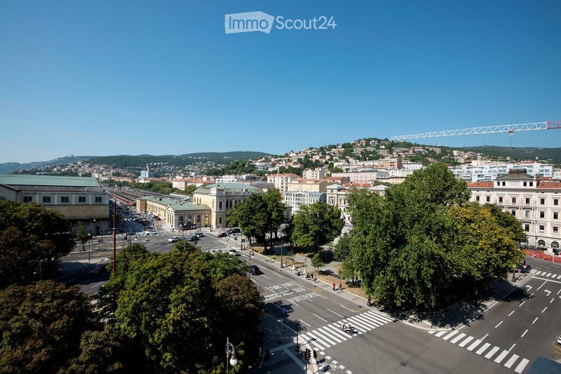 A panoramic view of a cityscape with buildings, roads, and greenery, featuring a clear blue sky and distant hills.