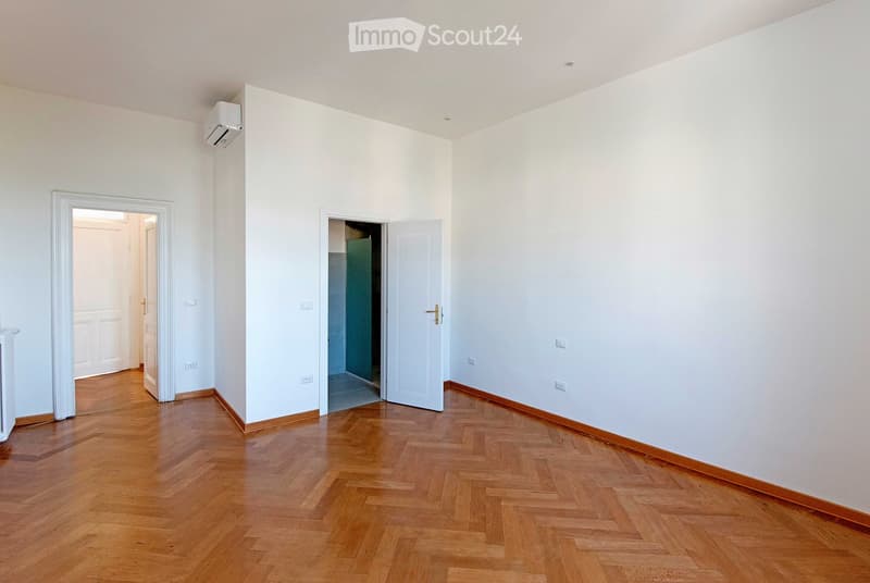 Empty room with light-colored walls, wooden herringbone flooring, and two open doors leading to other spaces.