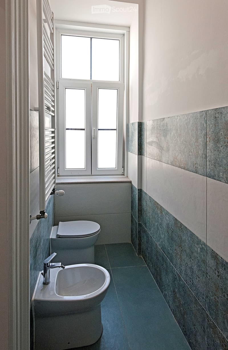 Modern bathroom with a toilet, sink, and faucet, featuring grey tiles and a window with white frames.