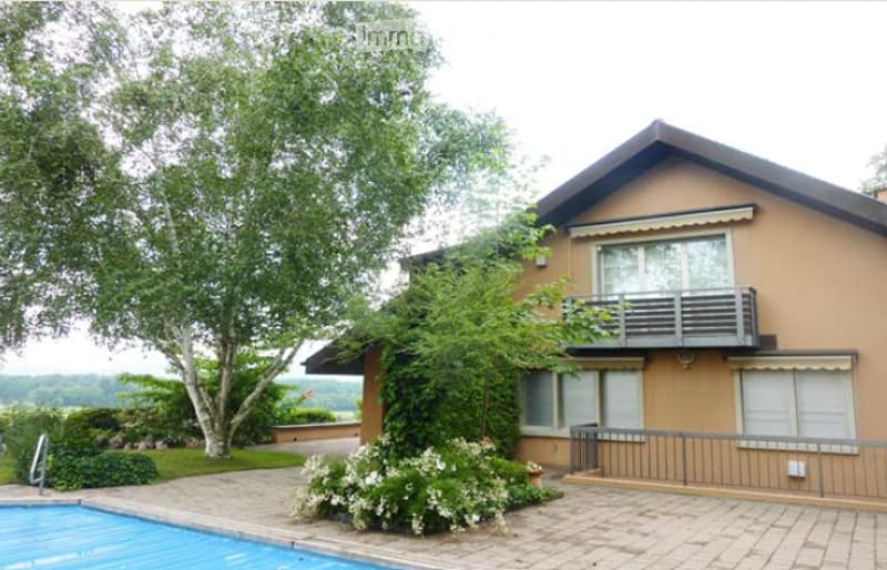 Two-story house with beige exterior, white-framed windows, and a balcony. Features a swimming pool in the front yard, surrounded by greenery and a paved area.