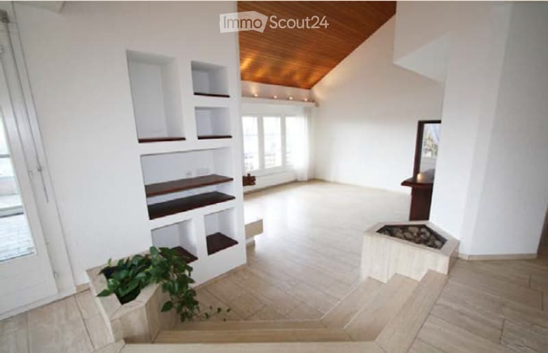 Bright, open room with white walls, wooden ceiling beams, built-in shelving units, and a small fireplace. The space is completely unfurnished, with large windows and a plant in the corner.