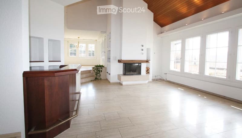 Bright, open hallway with white walls, large windows, a fireplace, and a wooden reception desk. The floor is tiled, and the ceiling has recessed lighting.
