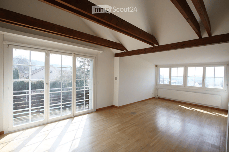 Bright, empty room with wooden flooring, exposed wooden beams, large windows, and a sliding glass door leading to a balcony with a view.