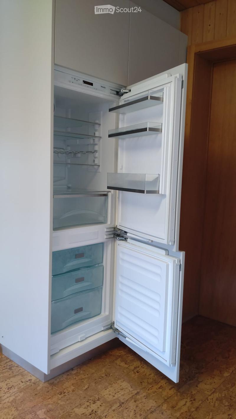A modern white refrigerator with multiple compartments and shelves, fully open, showing its interior. The kitchen has a wooden floor and light-colored cabinets.