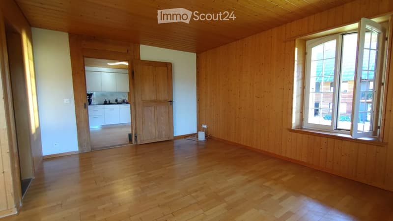 Empty room with wooden walls, wooden floor, large window, and a doorway leading to a small kitchen area.