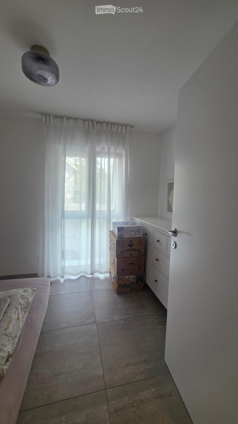 A furnished bedroom with a bed, a drawer unit, and a fan. The room has tiled flooring and white walls, with a window covered by sheer curtains.
