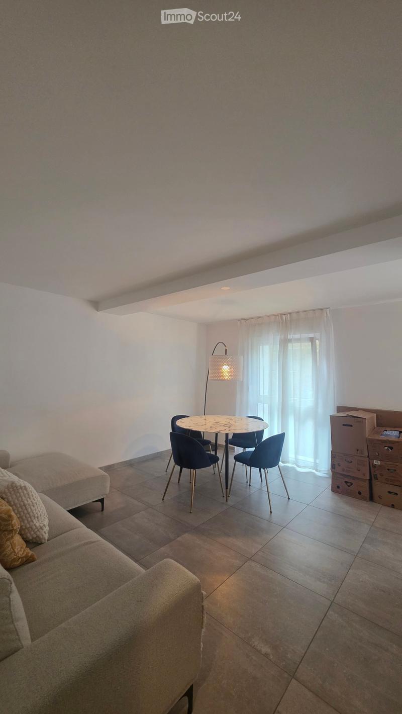 Living room with a beige sofa, a round wooden table with four blue chairs, a floor lamp, and several cardboard boxes stacked against the wall.