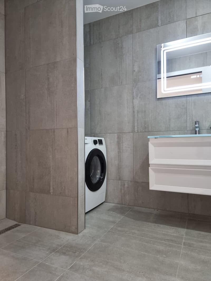 Modern bathroom with grey tiled walls, a white sink with a mirror, and a private washing machine.