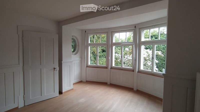 Empty room with light wooden flooring, white walls, multiple large windows, and a white door.