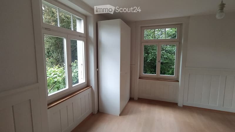 Empty room with wooden flooring, white walls, and large windows showing greenery outside. No furniture or equipment present.