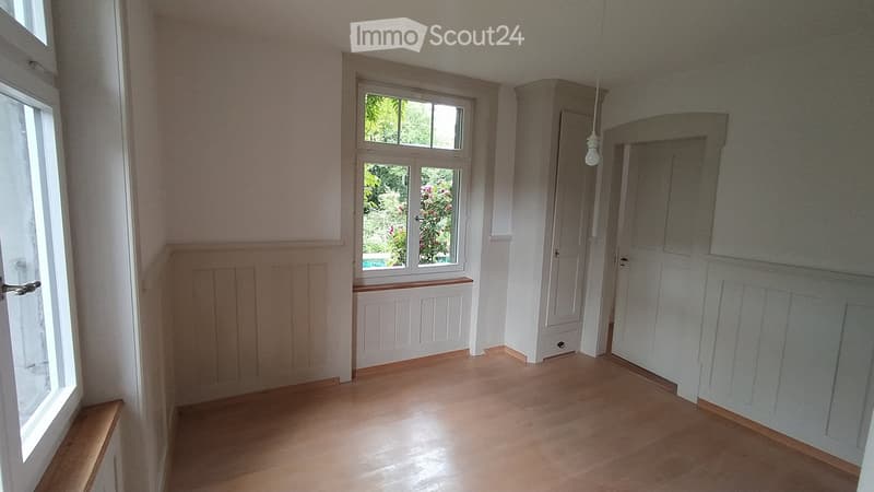 Empty hallway with wooden flooring, white walls, and multiple doors leading to other rooms. Natural light streams in from a window showing greenery outside.