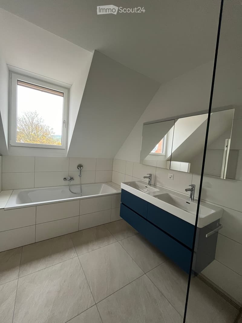 Modern bathroom with a bathtub, double sink, and large mirror, featuring light grey tiles and a skylight window.