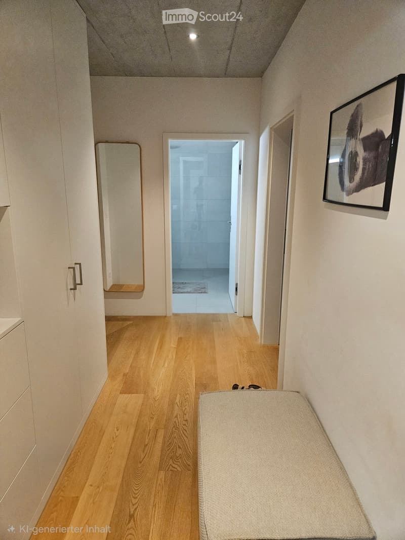 A clean, modern hallway with light wooden flooring, white walls, and a small bench. A framed picture hangs on the wall, and a mirror is mounted on the left side. An open doorway leads to another room.