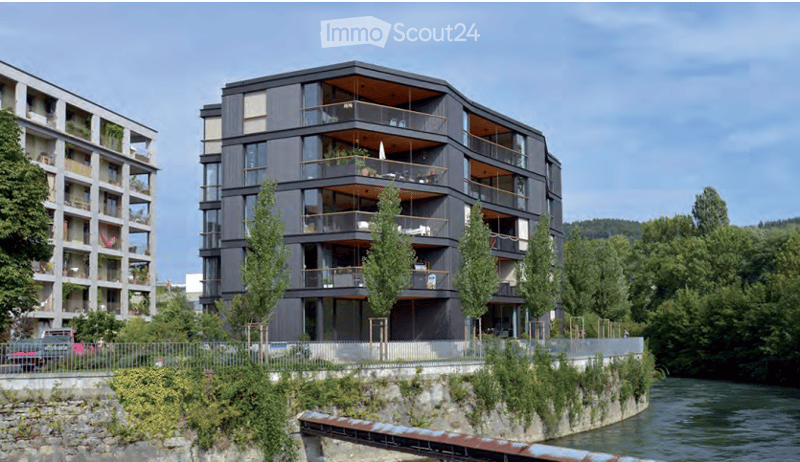 Modern multi-story building with balconies overlooking a river, surrounded by greenery.